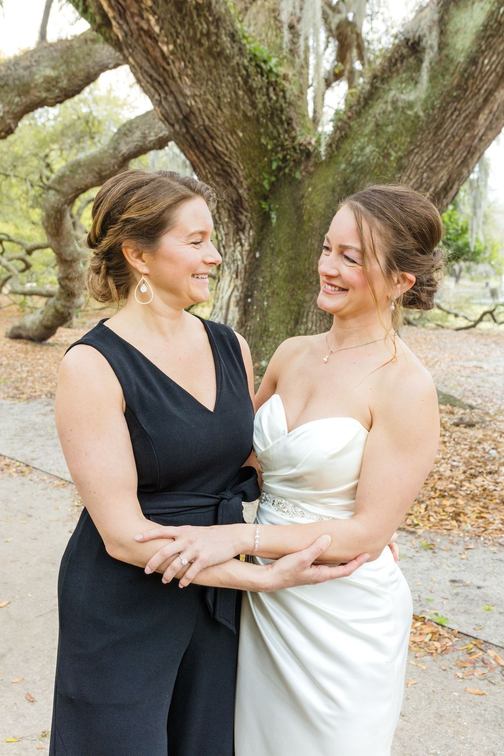 Two women in formal attire share laughs and embraces during an outdoor photoshoot.