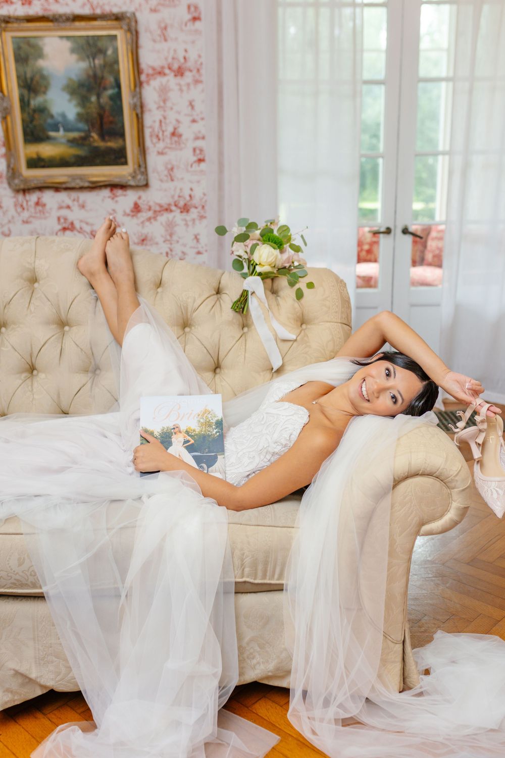 A bride in a white wedding gown reclines elegantly on a tufted cream chaise lounge.