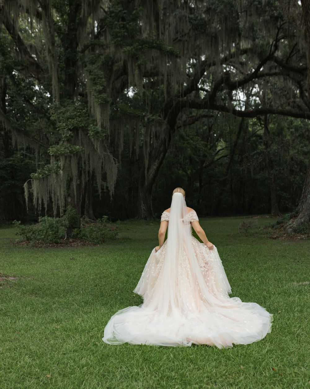 A bride in a blush wedding gown poses in a mossy oak garden setting.