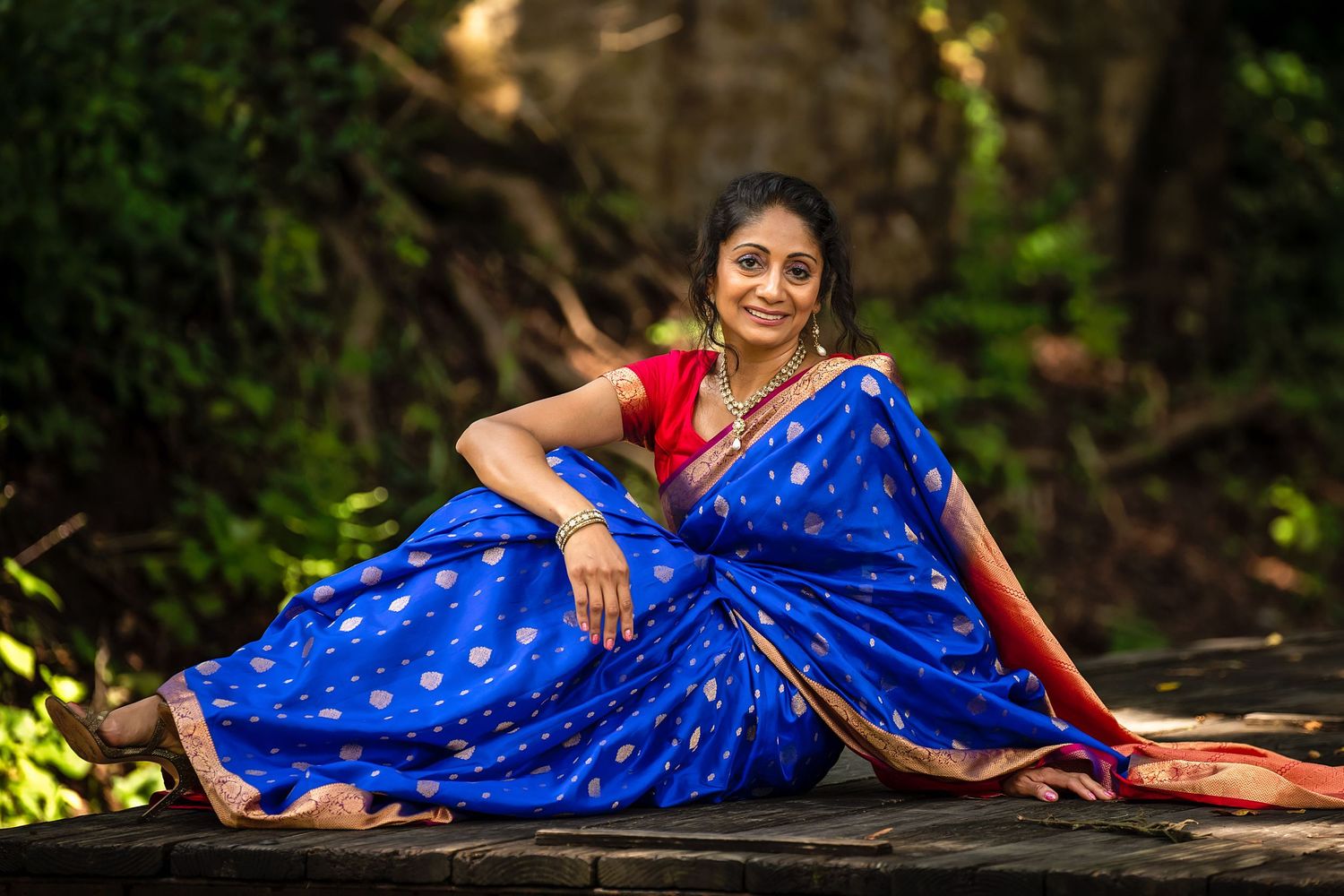 A person in a bright royal blue and red traditional saree poses elegantly outdoors against a natural backdrop.