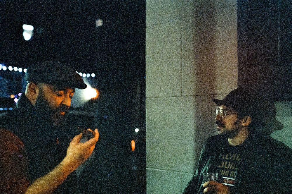 Two figures in dark clothing and hats stand in conversation at night.
