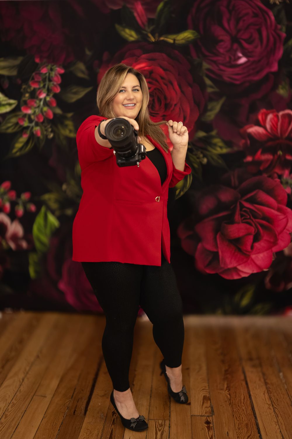 Professional photographer in a red blazer holding camera poses against floral backdrop with large red roses on hardwood floor.