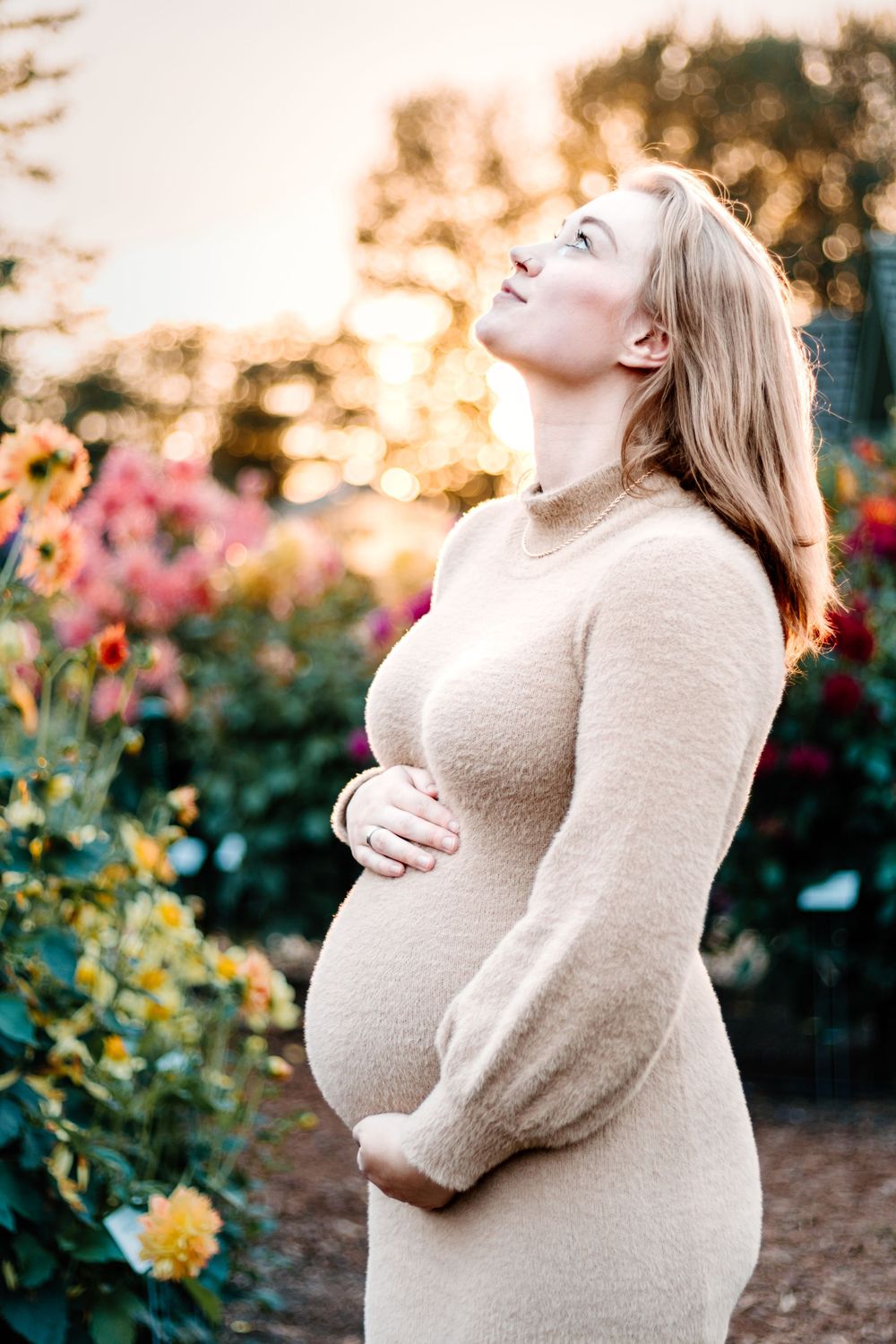 Expectant mother in beige sweater poses in garden during golden hour.