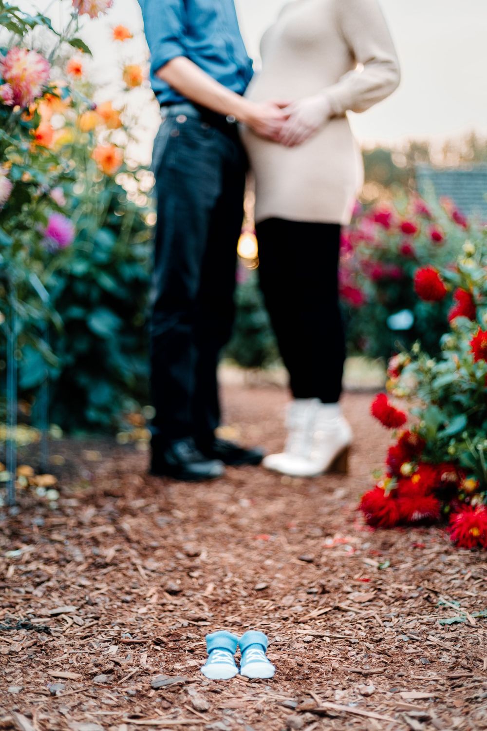 Baby shoes placed in foreground with couple standing among colorful garden flowers.