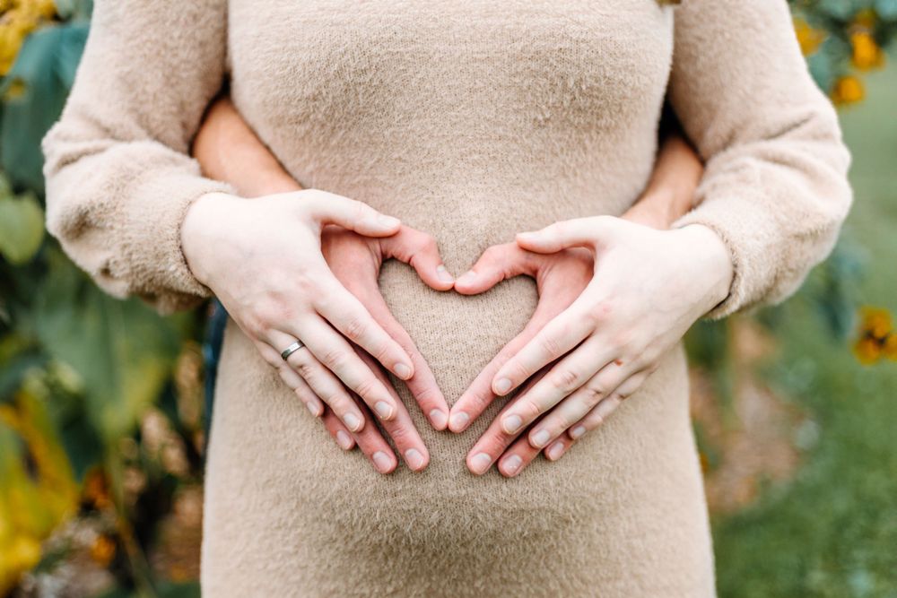 Hands form heart shape over pregnant belly against garden background.