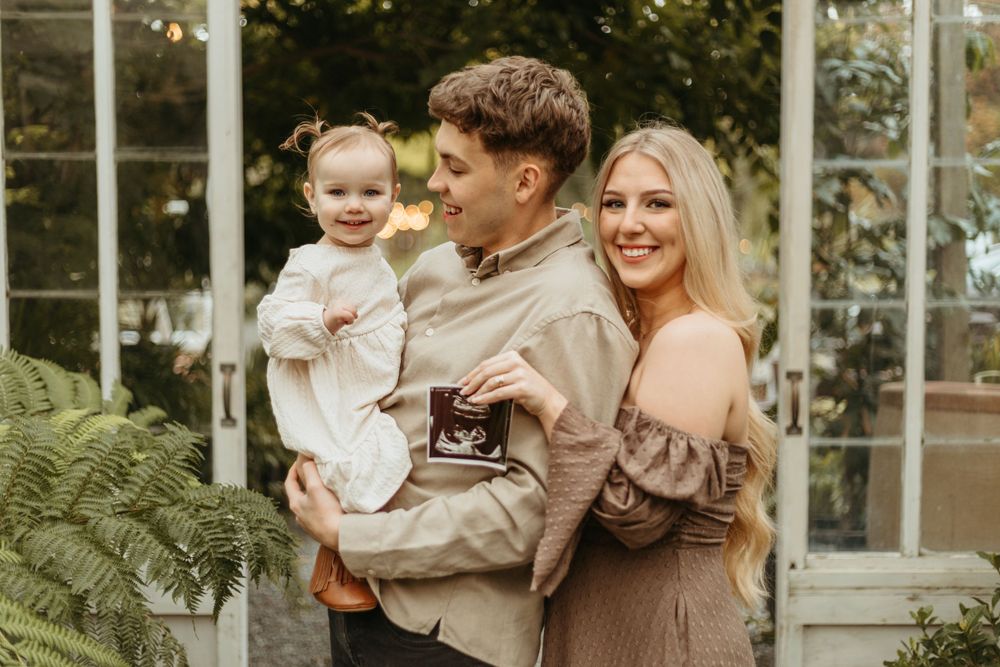 Family of three poses together inside a greenhouse with ferns and glass walls in the background.