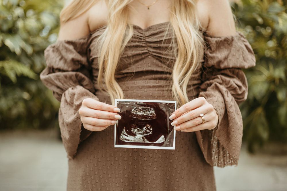 Hands holding an ultrasound photo against a brown dress in a garden setting.