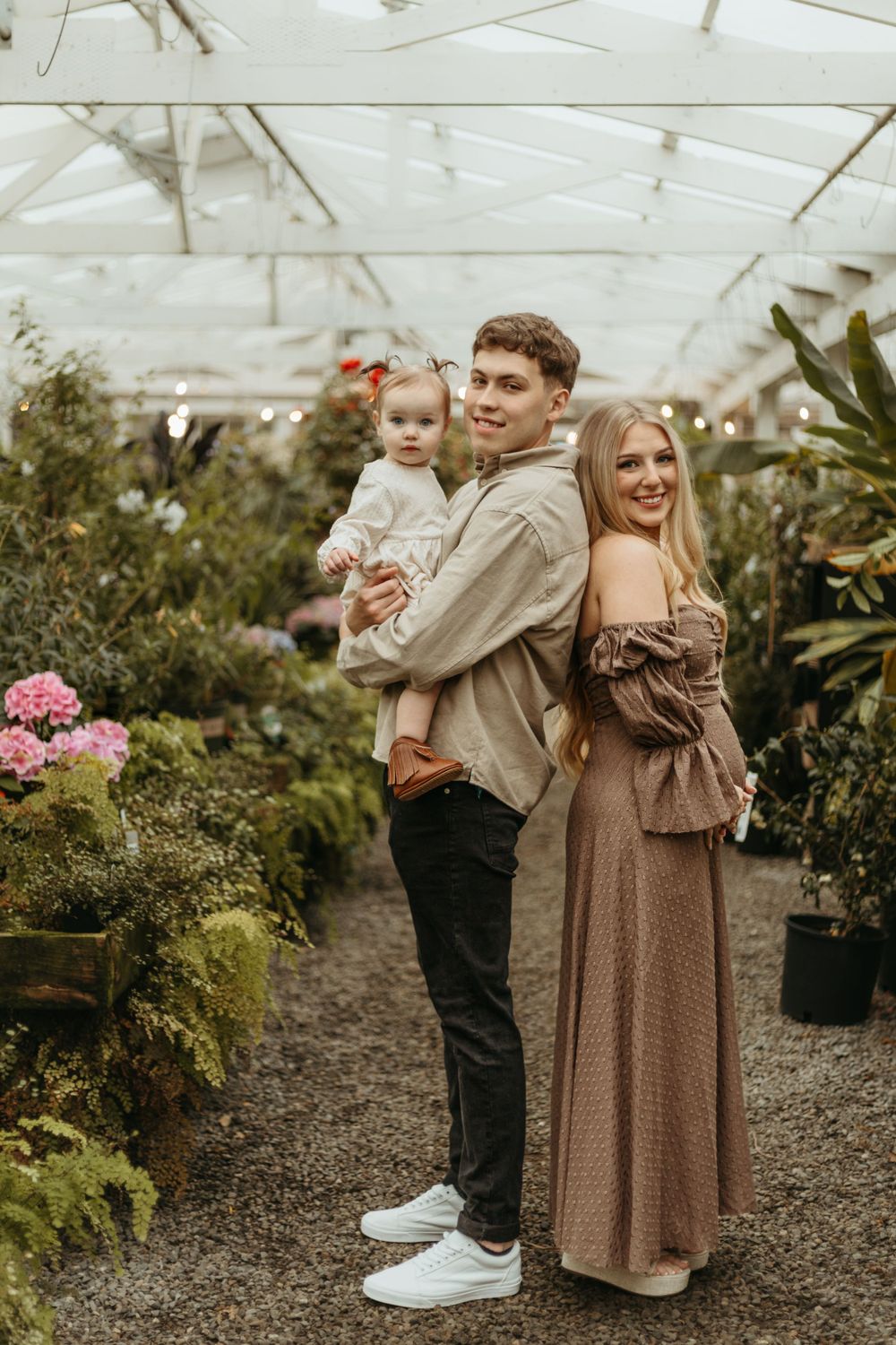 A couple and a small child pose together in a greenhouse filled with lush plants and pink flowers.