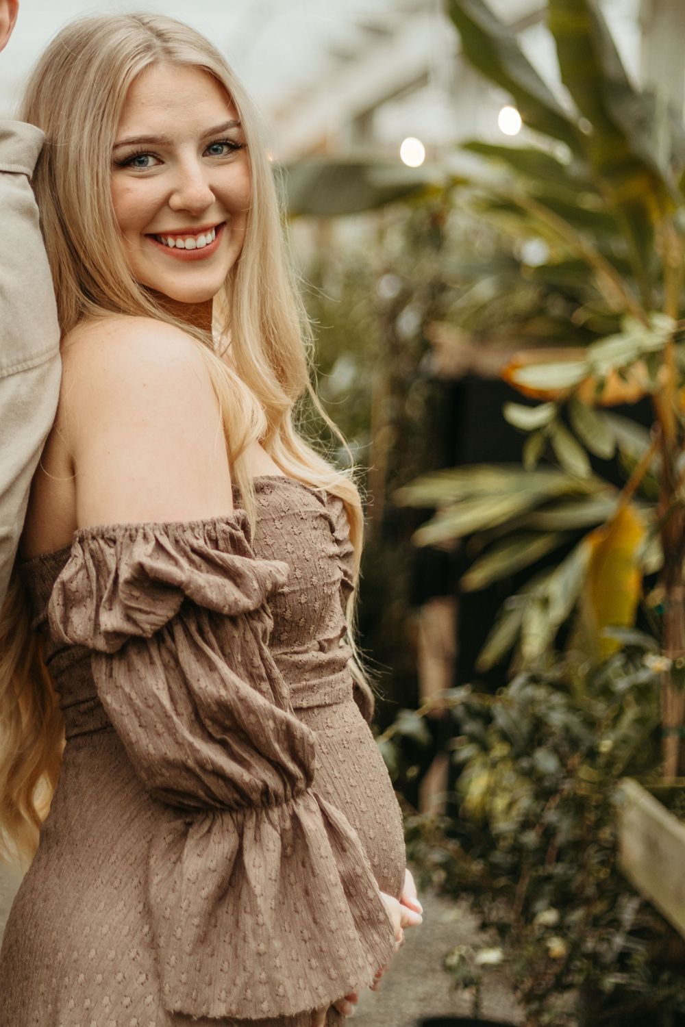 Woman in a brown off-shoulder dress smiles while standing among greenhouse plants.