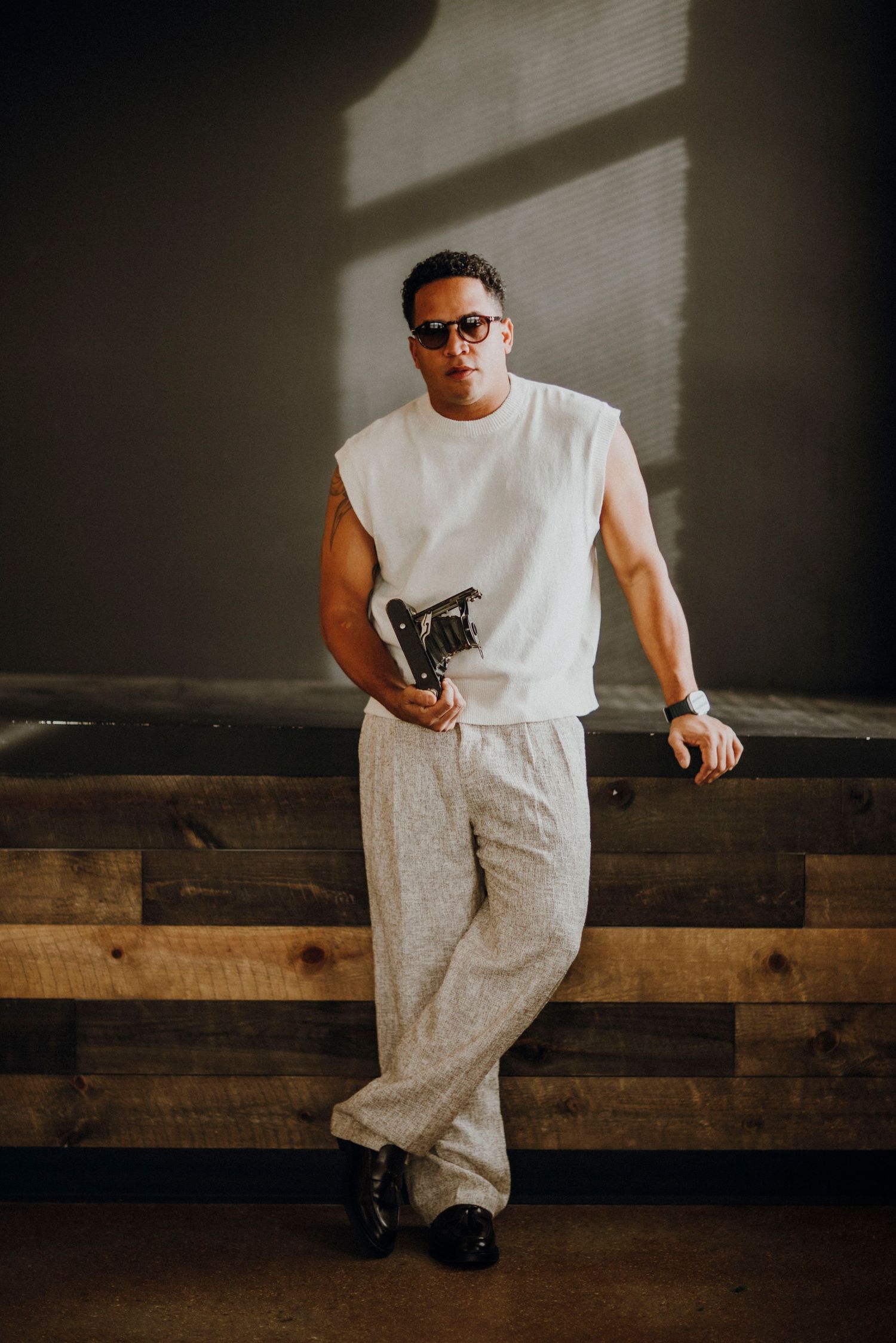 Casual portrait against wooden wall wearing white sleeveless shirt and grey pants.