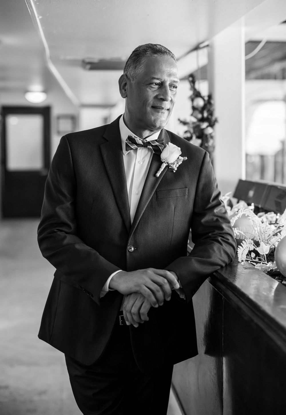 A distinguished gentleman in a black tuxedo and bowtie poses in a hallway on wedding day.