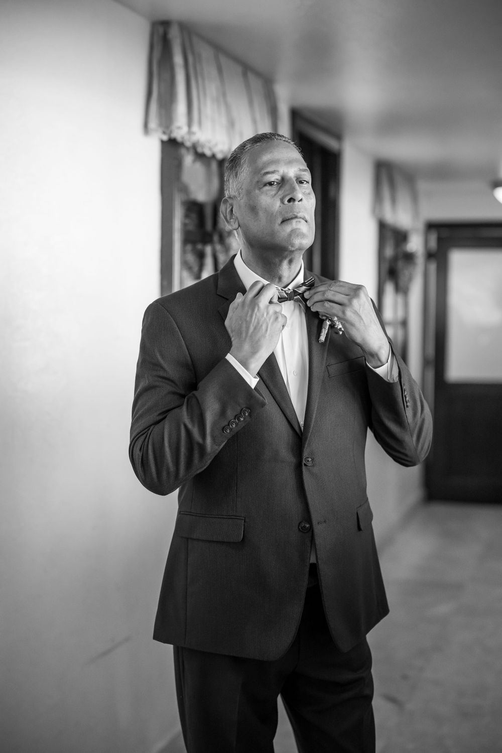 A groom adjusts his bow tie while preparing for his wedding day in a classic black suit.