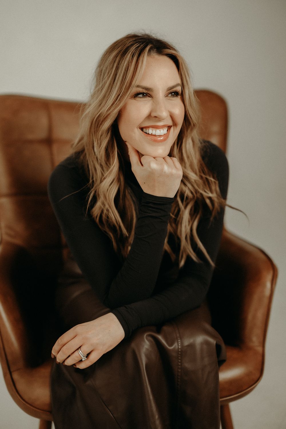 Person in black sweater sitting in brown leather chair with relaxed, cheerful expression.