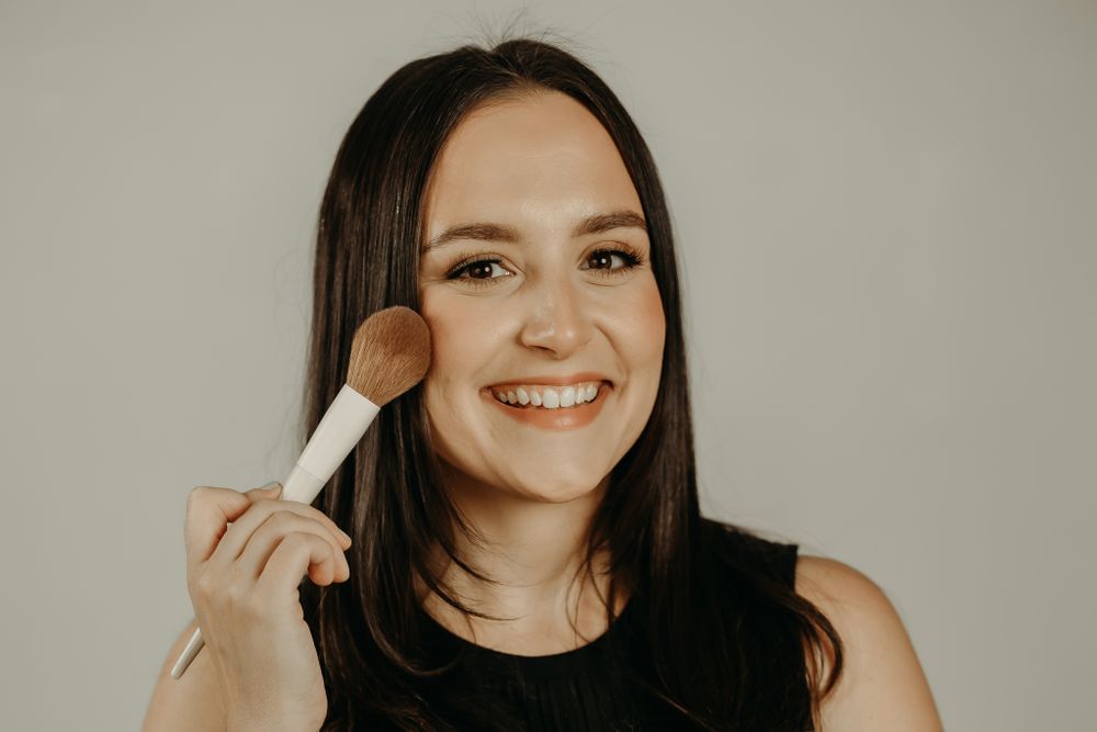 Person holding a makeup brush against their cheek while smiling at the camera.