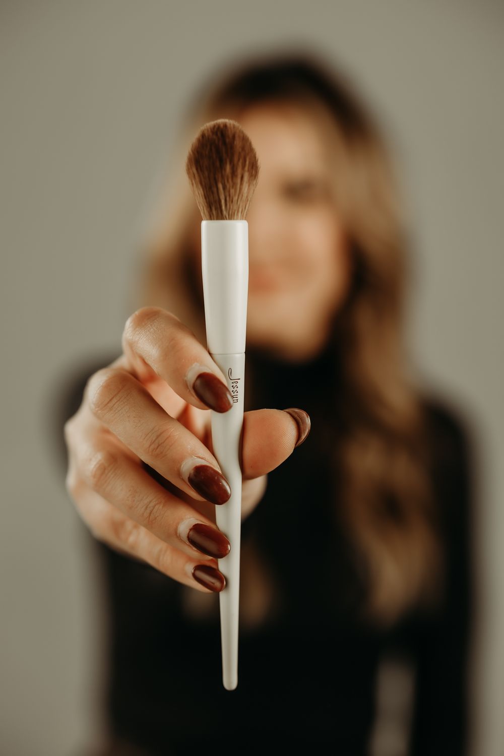 Close-up of a hand with burgundy nails holding a white makeup brush against a blurred background.