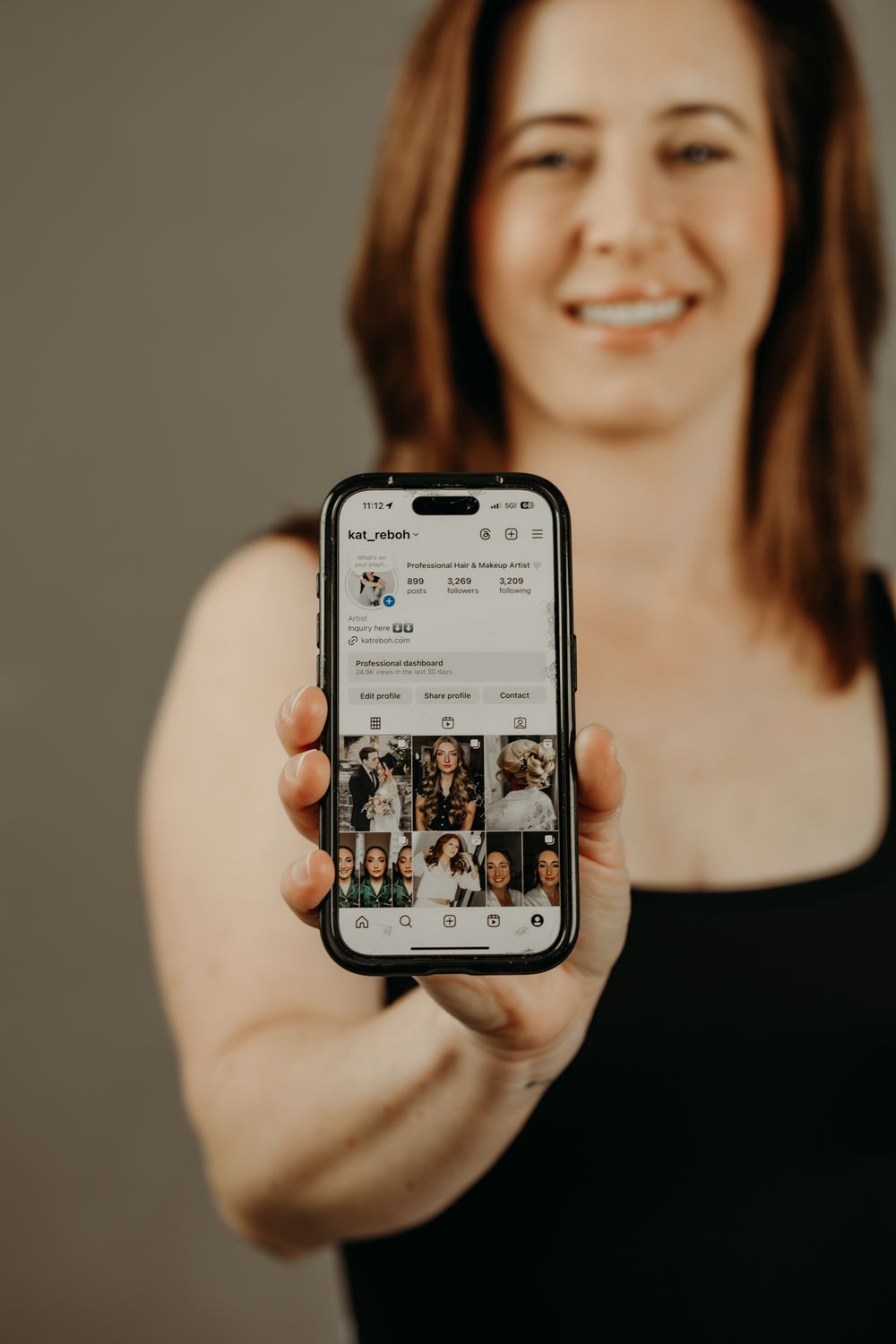 Hand holding iPhone displaying Instagram profile grid against neutral background.