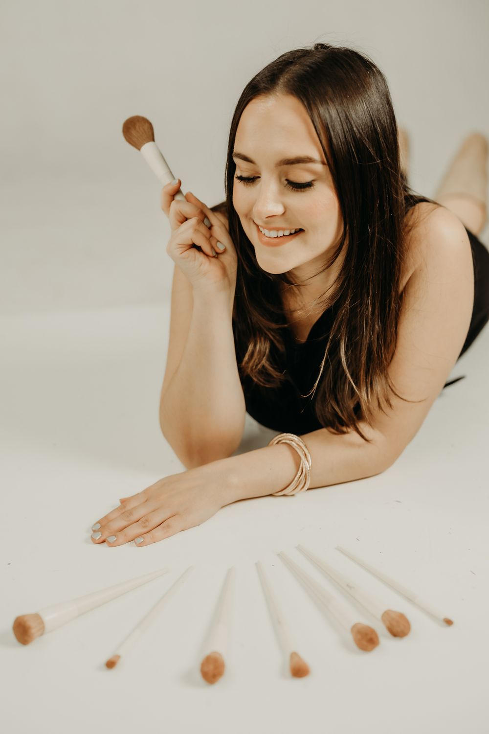 Person lying down arranging makeup brushes in fan pattern on white surface.