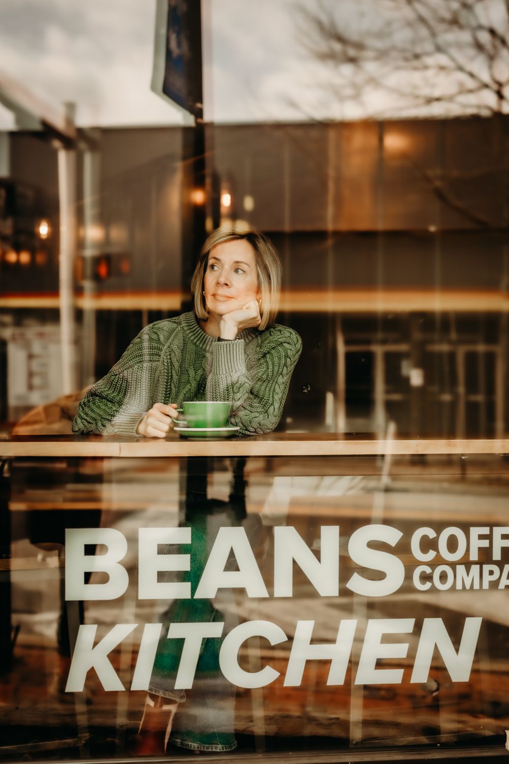 A person in a green sweater sits by a window at Beans Coffee Company Kitchen with a green coffee cup.
