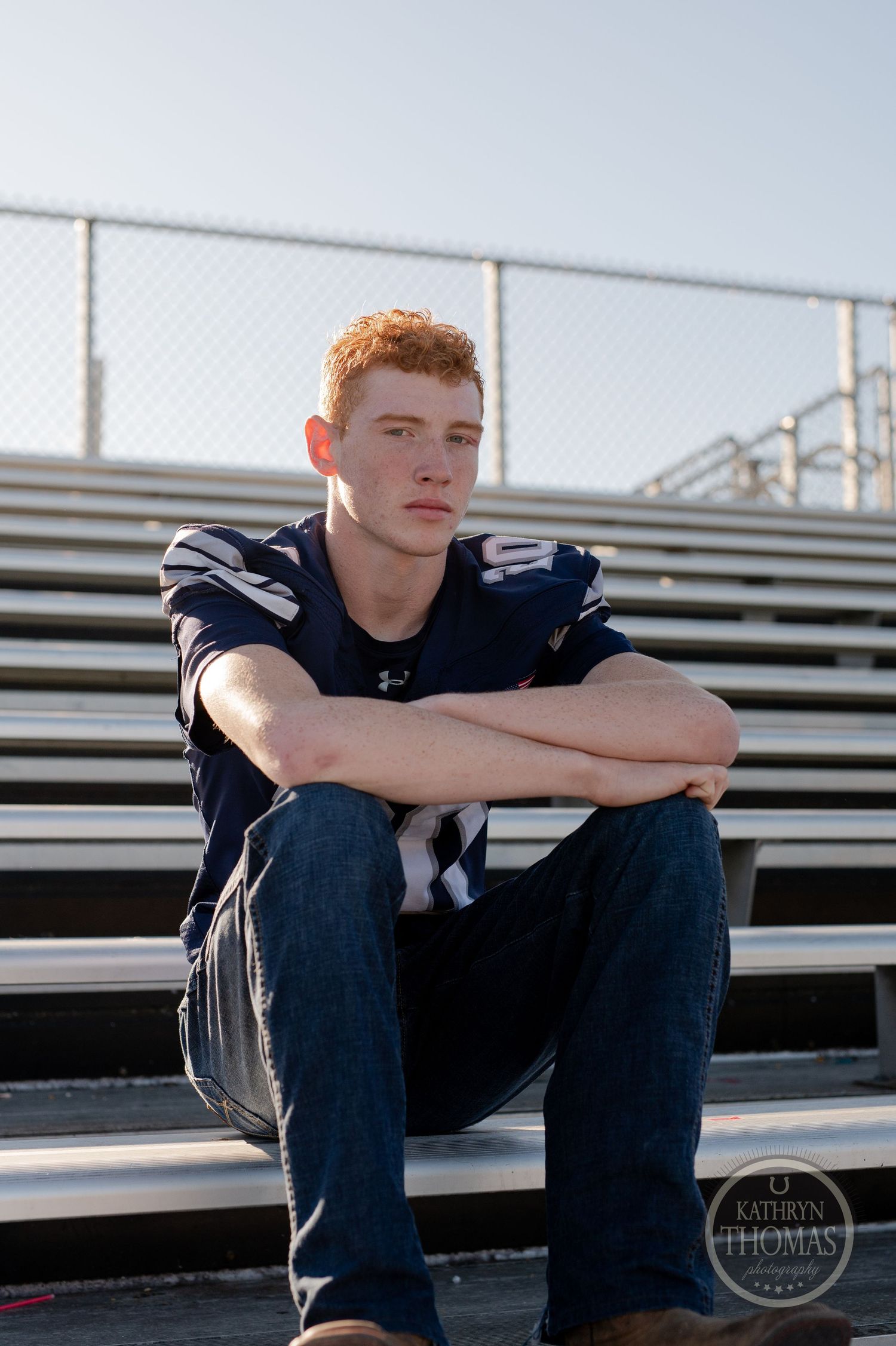 An athlete in a navy football jersey sits contemplatively on metal bleacher seats.