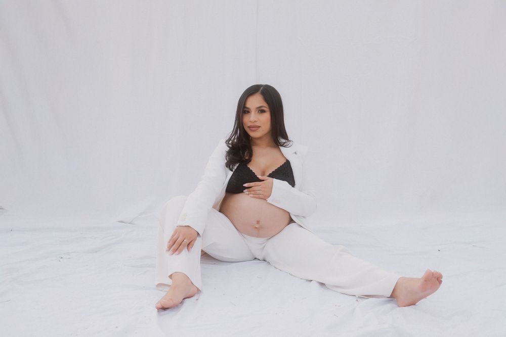 Elegant maternity pose on white backdrop wearing white pants and blazer.