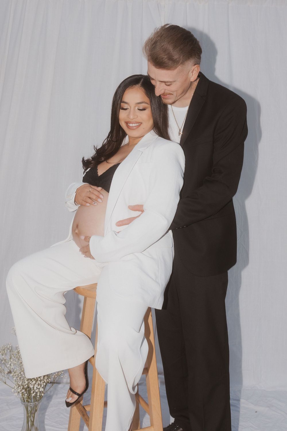 Studio maternity portrait featuring couple in formal attire against white backdrop.