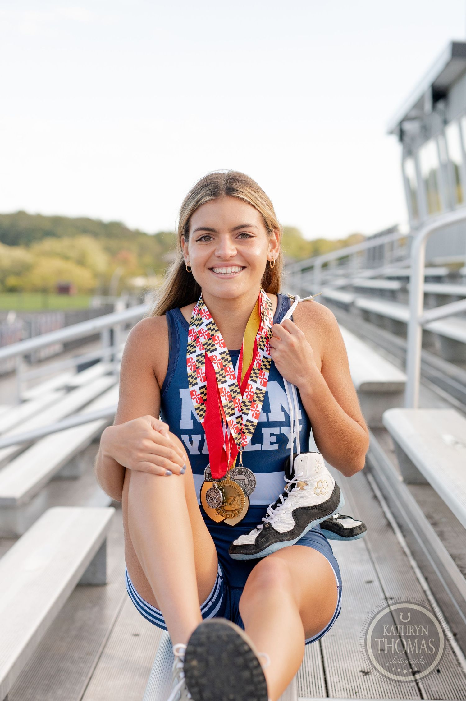 Student athlete wearing blue uniform showing off medals while sitting on metal bleachers.