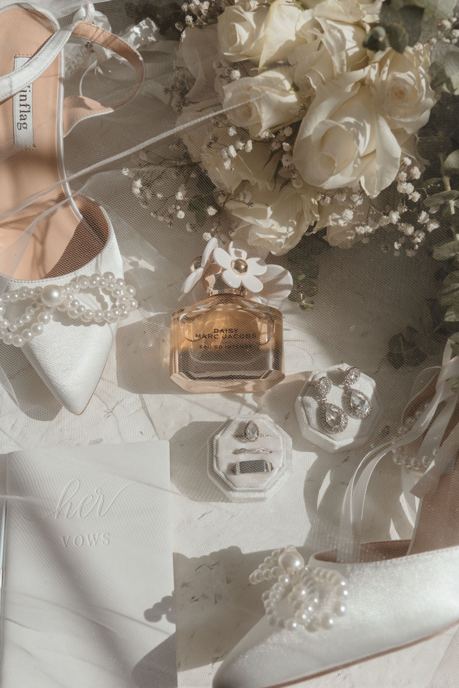 Wedding details flatlay with perfume bottle, shoes, and white flowers in soft lighting.