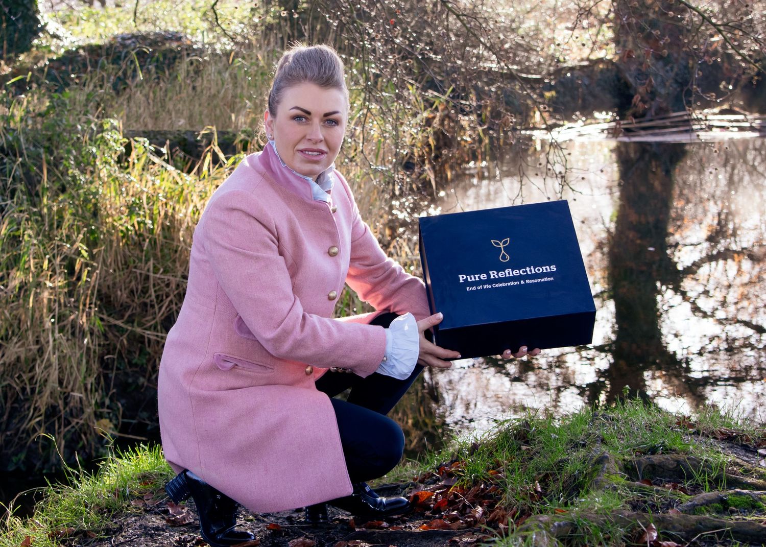 Person in pink coat examining navy blue box by water's edge.