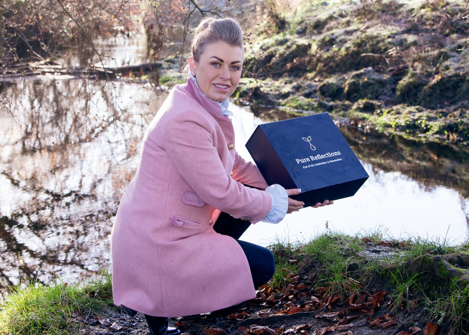 Person in pink coat holding navy blue box outdoors in autumn setting.