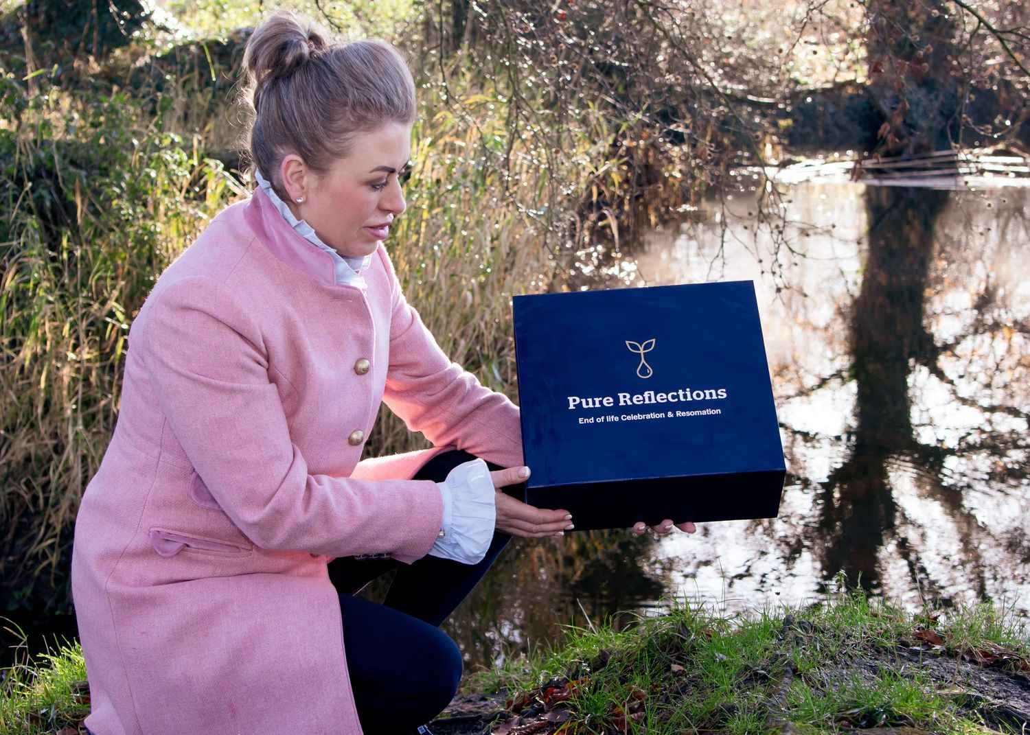 Person in pink sweater holding branded box near pond in natural setting.