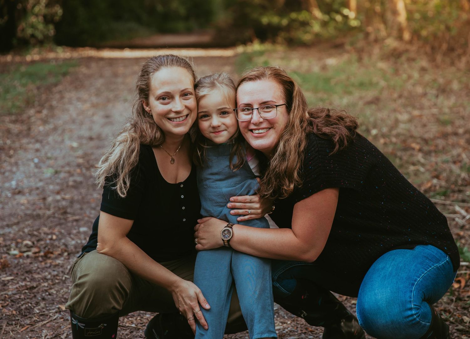 Three people kneeling closely together on a dirt path surrounded by trees at dusk.