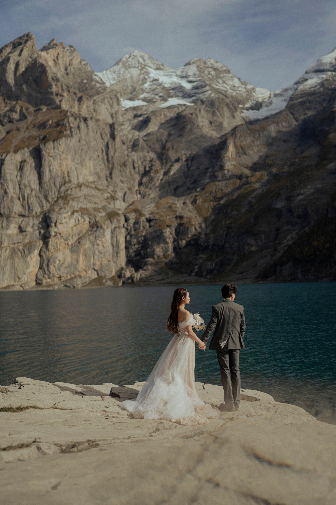 Newlyweds admiring mountain scenery by turquoise lake in dramatic landscape setting.