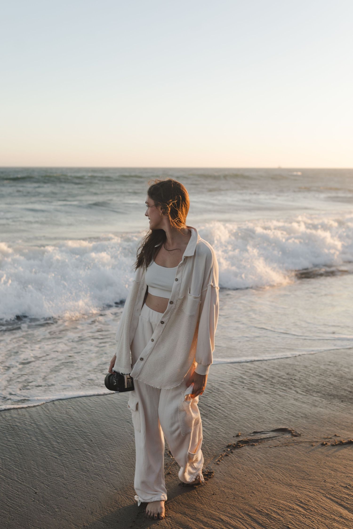 Person wearing white beach outfit standing on sandy shore at sunset with crashing ocean waves in background.