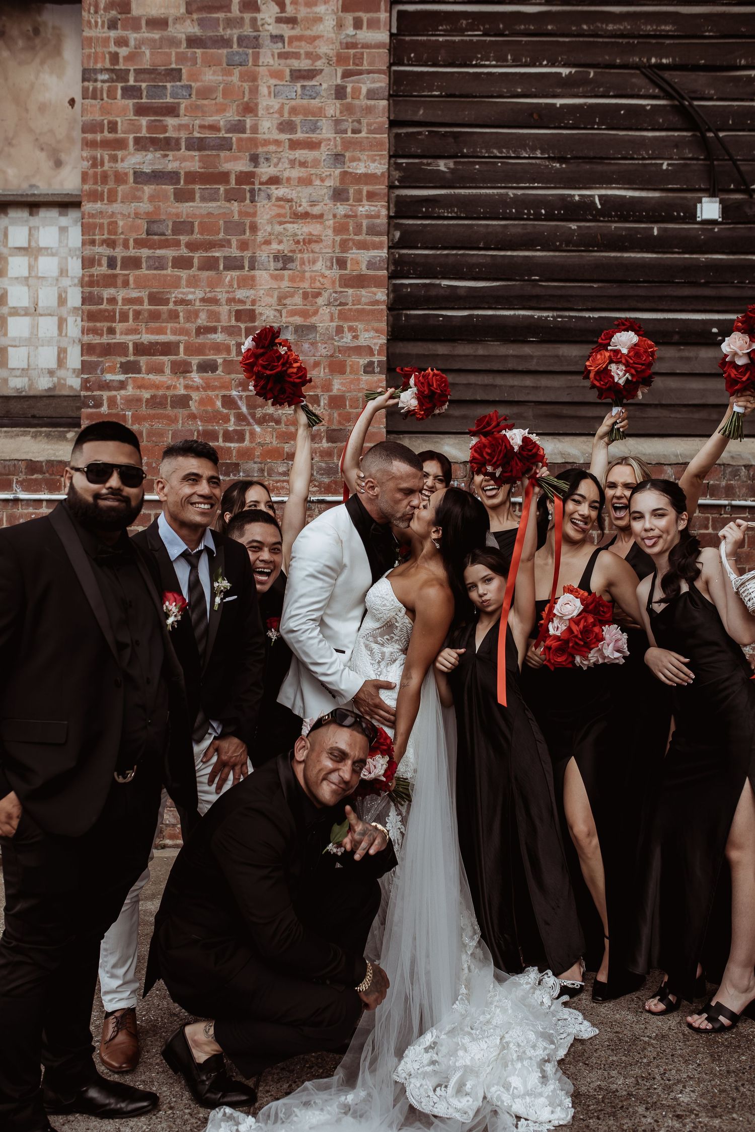 Wedding party in formal attire poses playfully against brick wall with red bouquets and black bridesmaids dresses. Brisbane Wedding