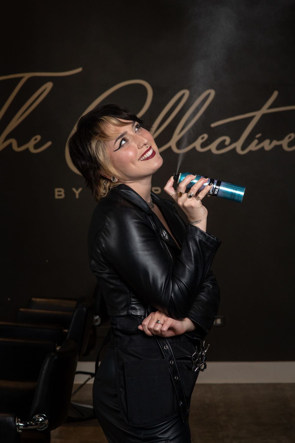 Woman in black leather jacket spraying hair product at The Collective salon, smiling confidently.