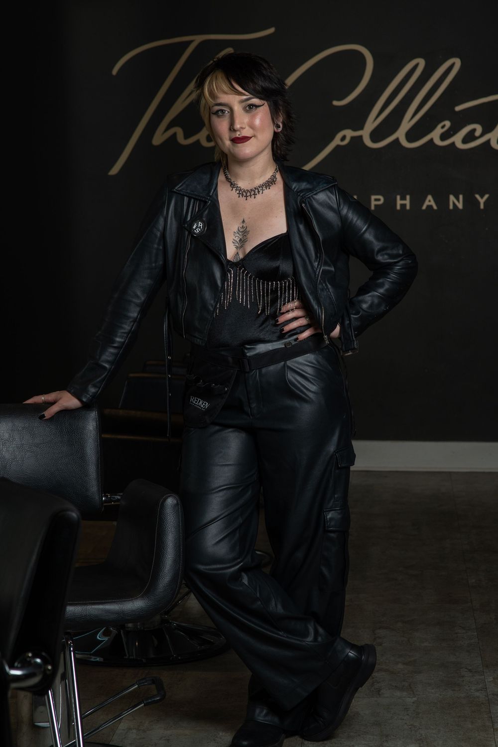 Woman in all-black leather outfit poses confidently in a dark salon studio with gold script signage behind her.