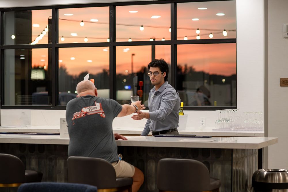 Hotel front desk staff member handing a drink to a guest at a modern check-in counter during sunset.