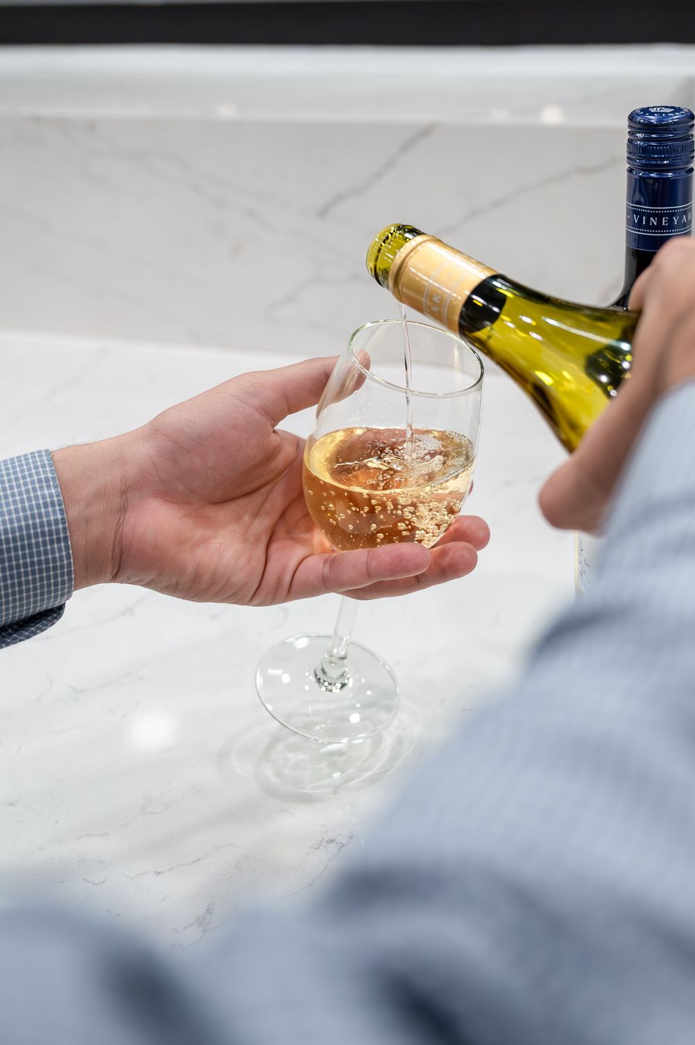 Person pouring white wine from a bottle into a stemless glass held over a marble surface.