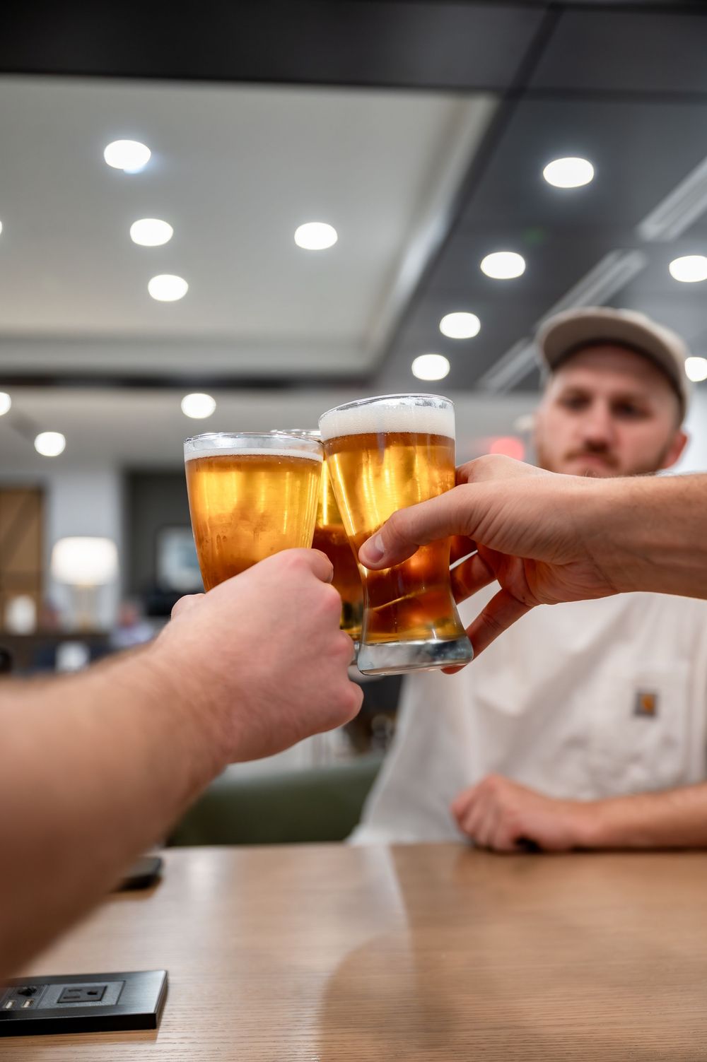 Two people toasting with full glasses of golden beer at a bar, with a man in a cap visible in the background.