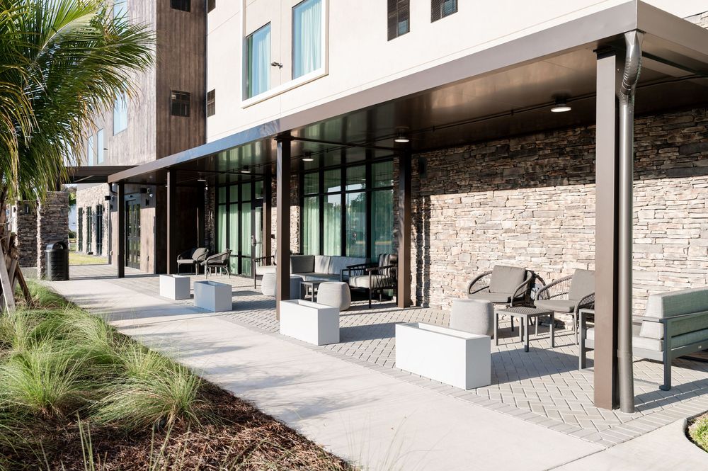 Modern hotel patio with stone walls, glass windows, outdoor seating, and landscaping on a sunny day.