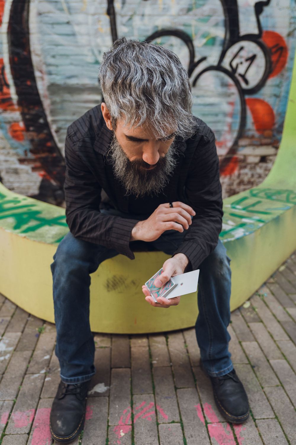 Bearded man with gray hair sits on a bench holding playing cards in front of a colorful graffiti wall.