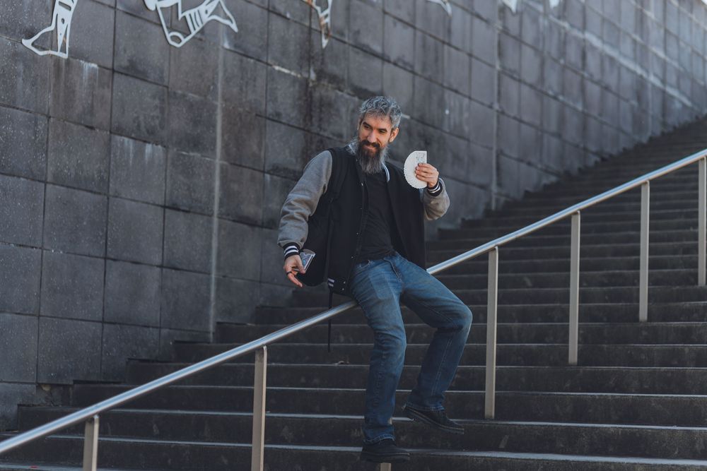 Man in black vest and jeans posing on outdoor stone stairs while holding papers near metal railing.
