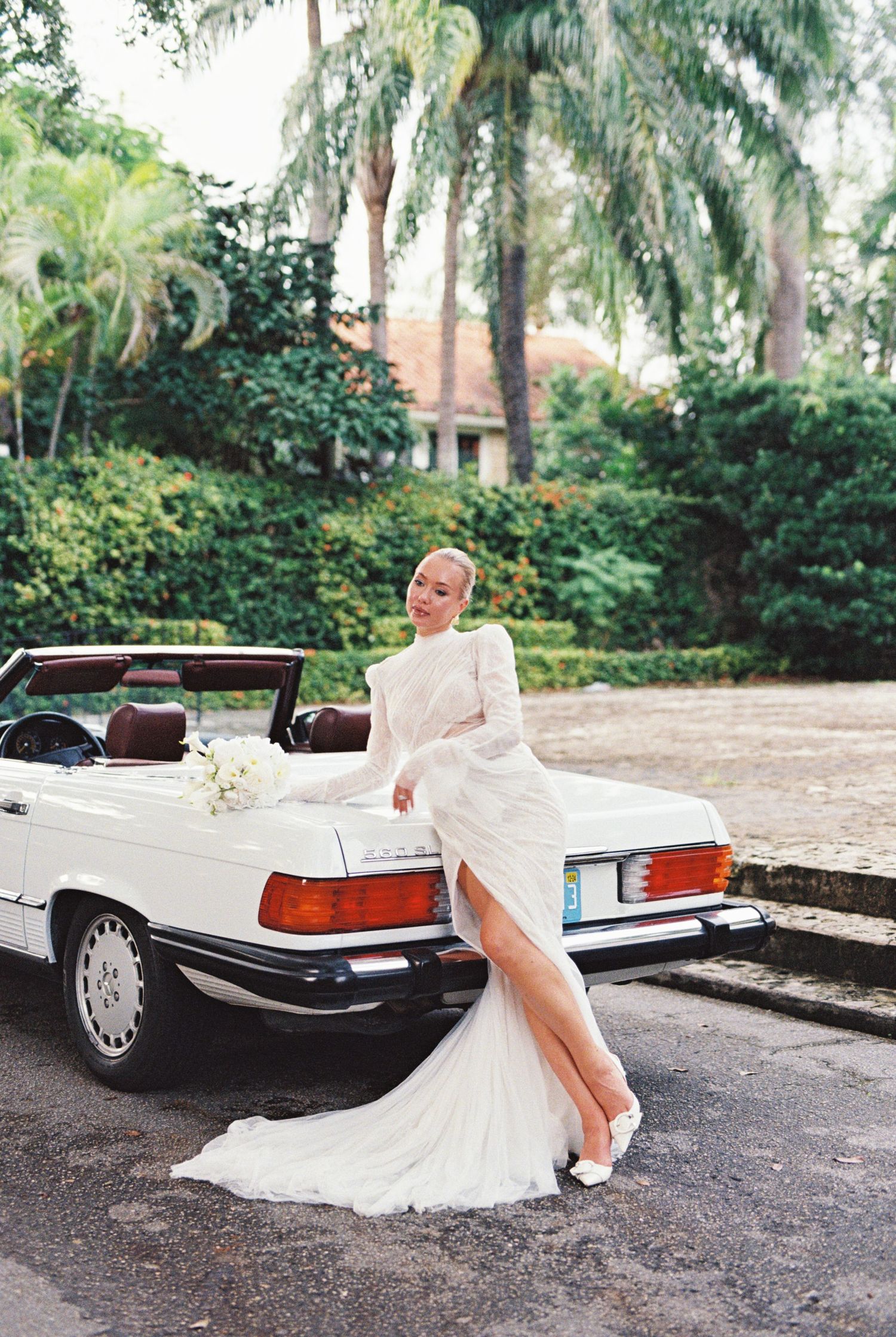 A luxurious white convertible Mercedes-Benz SL parked in a tropical garden setting with palm trees in the background.