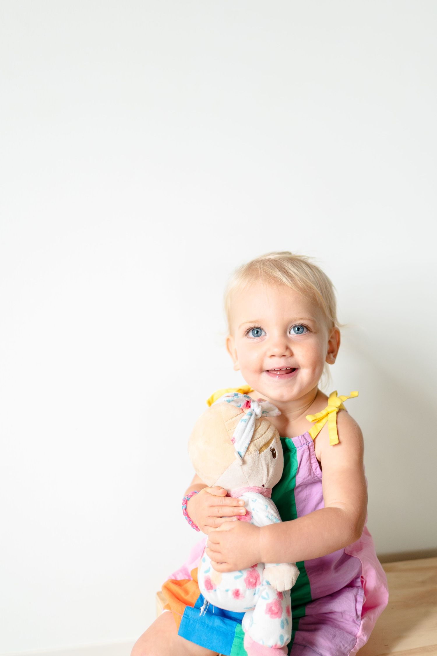 Happy toddler in pink dress hugging stuffed toy on plain white background.
