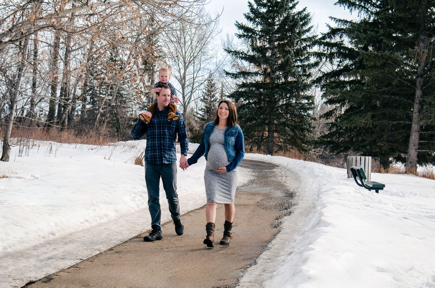A couple walks together holding hands on a snowy winter path surrounded by evergreen trees.