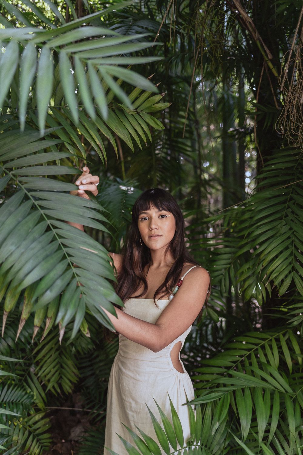 A person in a white dress poses elegantly among lush tropical palm fronds in a garden setting.