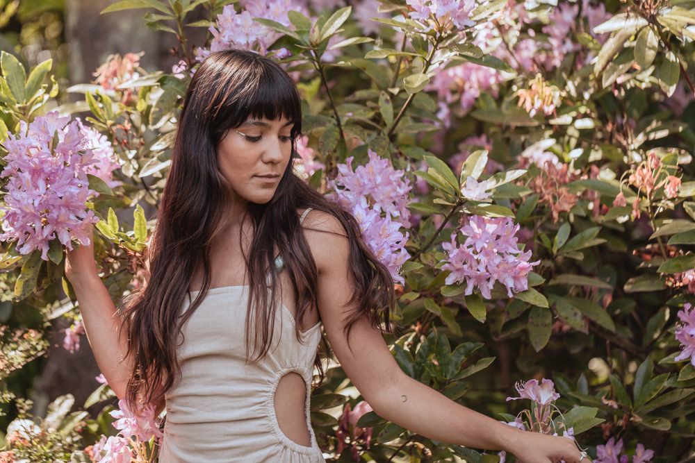 A figure in a white dress stands among blooming purple rhododendron flowers in a garden.