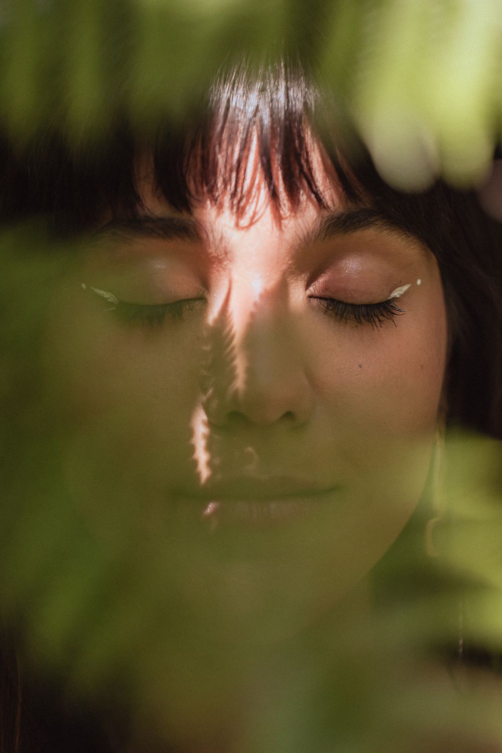 Close-up artistic portrait of someone with closed eyes and bangs in soft natural light with green bokeh effect.