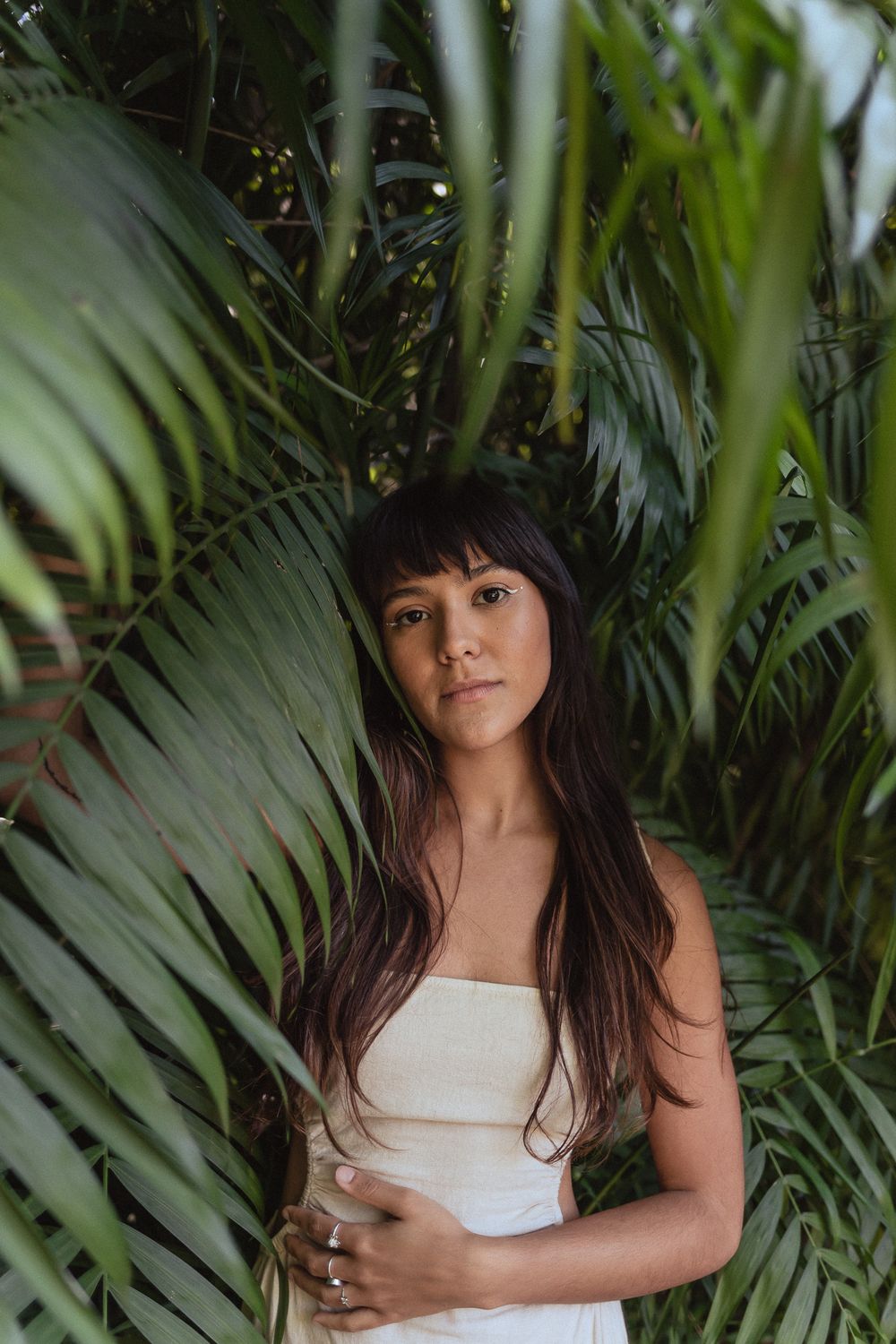 Person in white dress poses among tropical palm fronds in moody outdoor portrait.