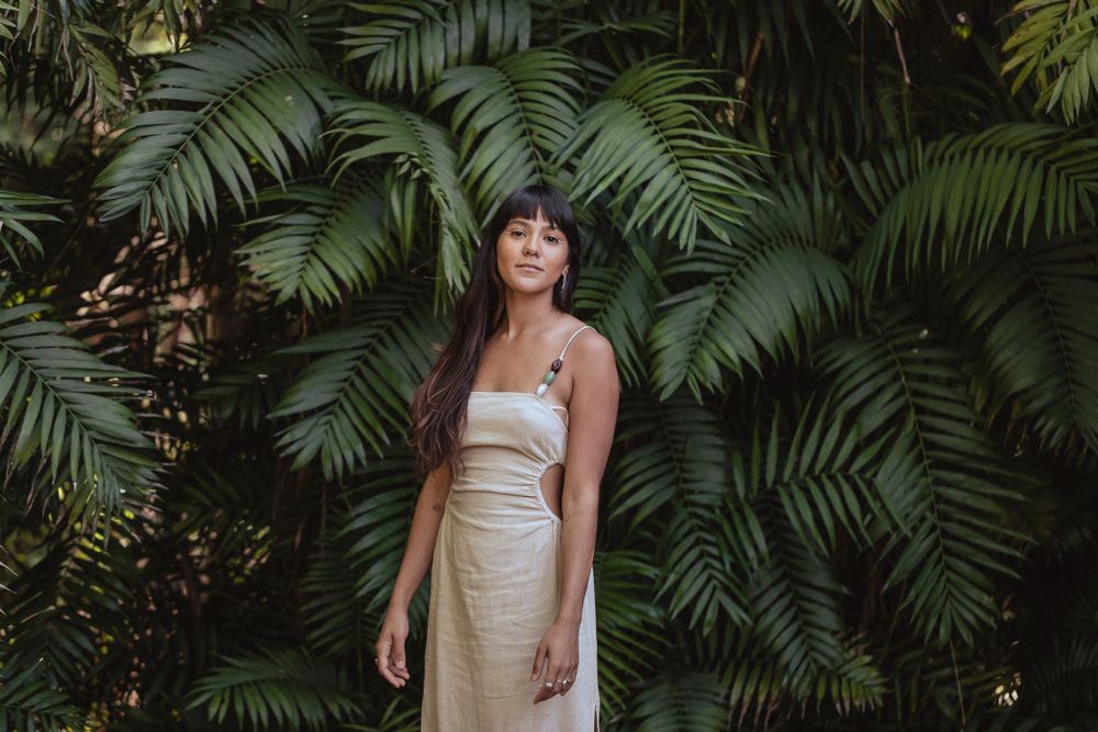 A person in a cream-colored dress poses against a backdrop of lush tropical palm fronds in a garden setting.